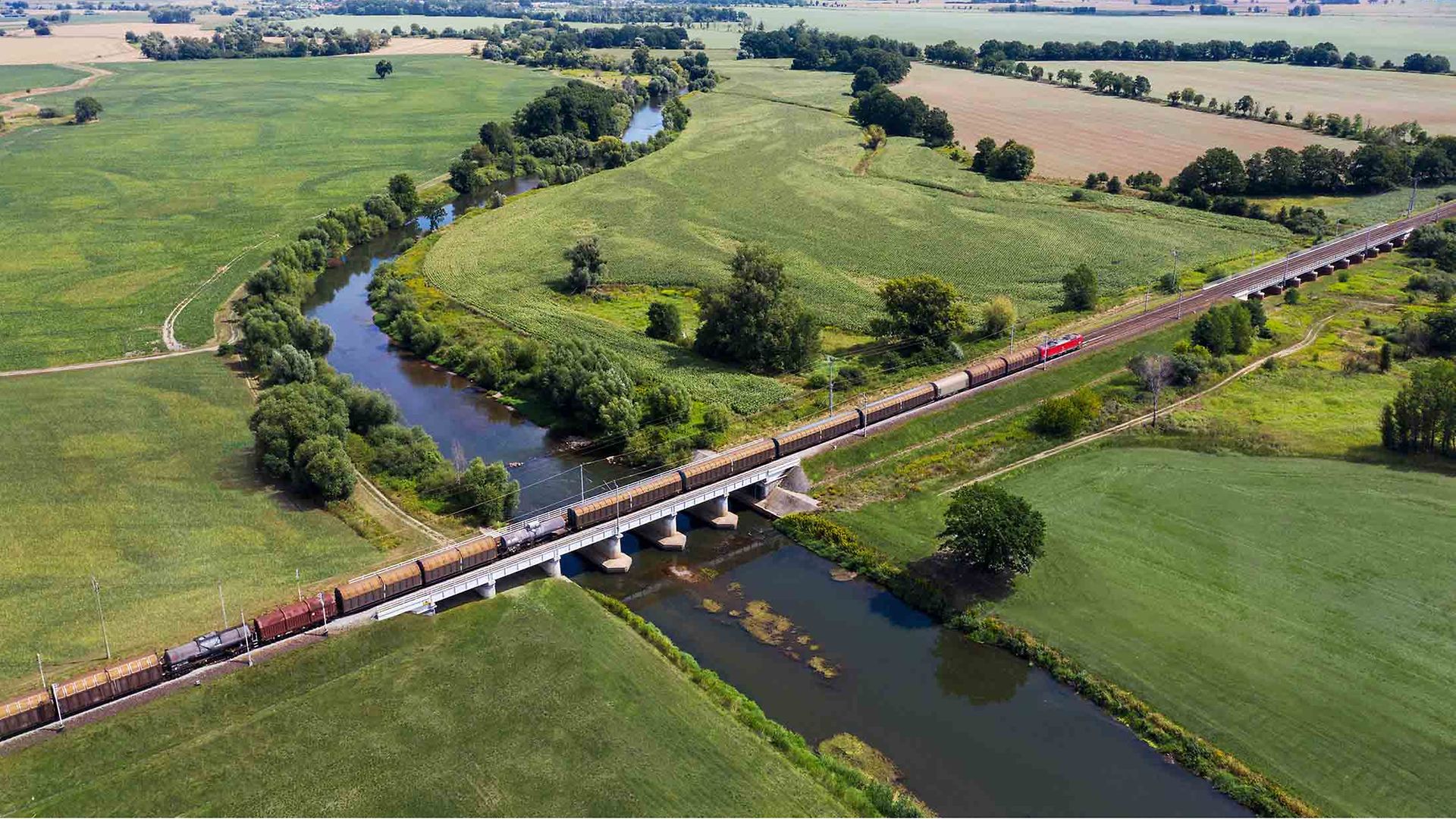 Güterzug fährt über einen Fluss inmitten grüner Landschaft.