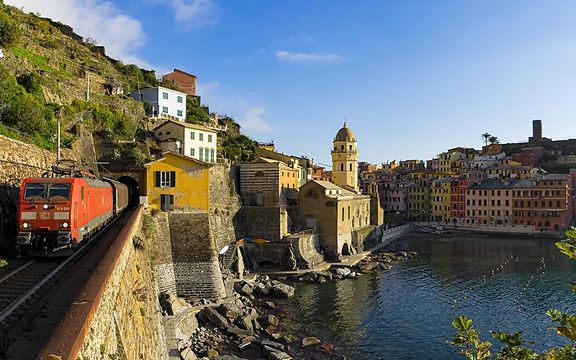 A DB Cargo freight train travels along a coast. Behind him you can see a small Italian town.