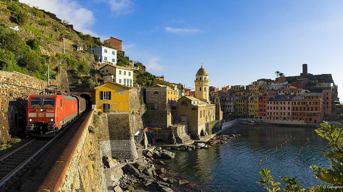 A DB Cargo freight train travels along a coast. Behind him you can see a small Italian town.