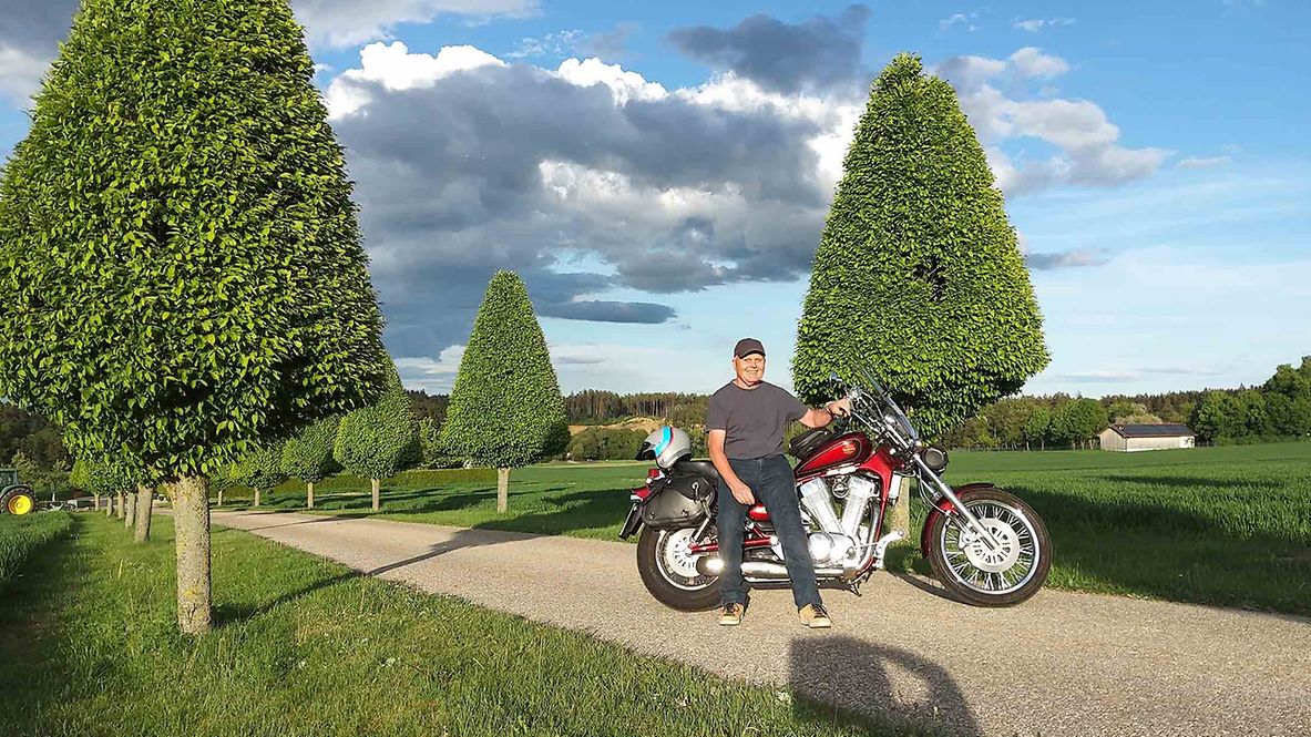 Ludwig Huber lehnt in einer Allee an einem Motorrad.