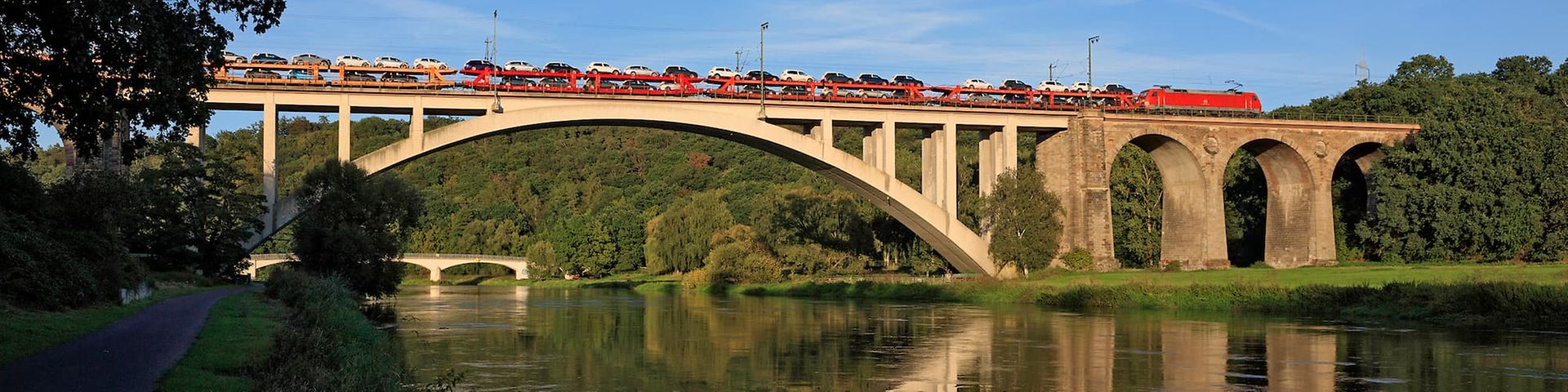 Ein Güterzug mit mehreren Güterwagen fährt unter blauem Himmel über eine Brücke.