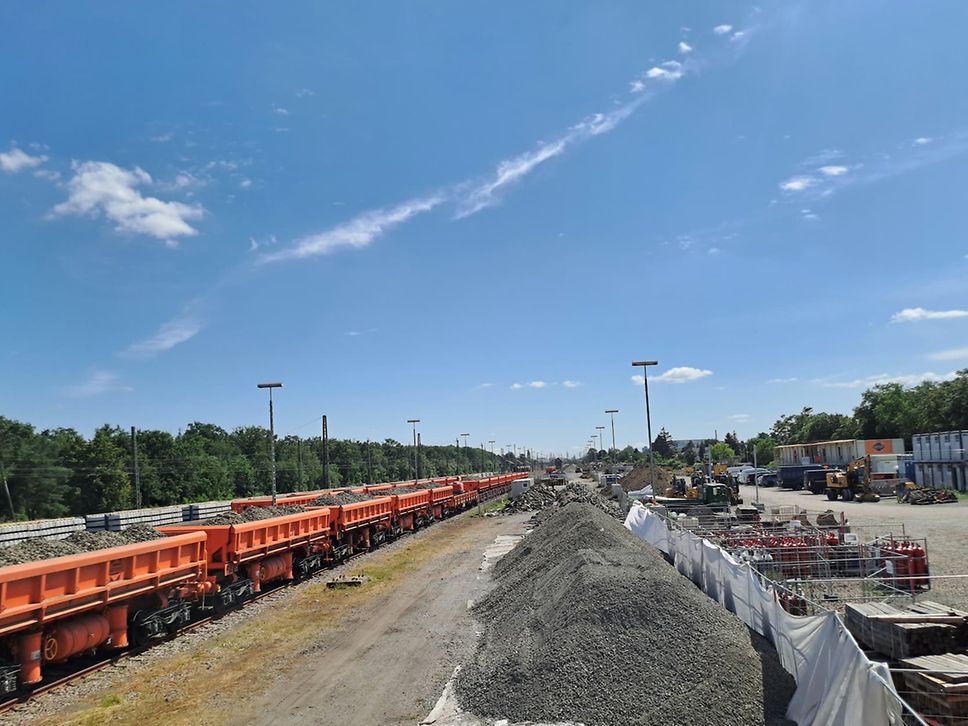 A freight train with orange wagons transports ballast. To the right you can see the construction site and piles of ballast.