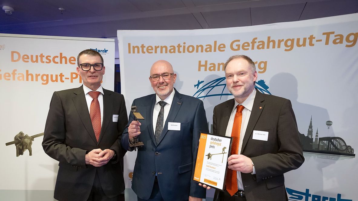 Drei Männer in Anzügen bei der Verleihung des Deutschen Gefahrgut-Preises 2025, einer hält die Trophäe, ein anderer die Urkunde.