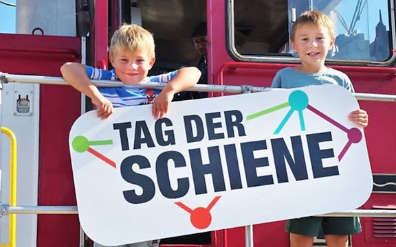 Kinder stehen auf einer Lok und halten zusammen ein Tag der Schiene Poster in den Händen