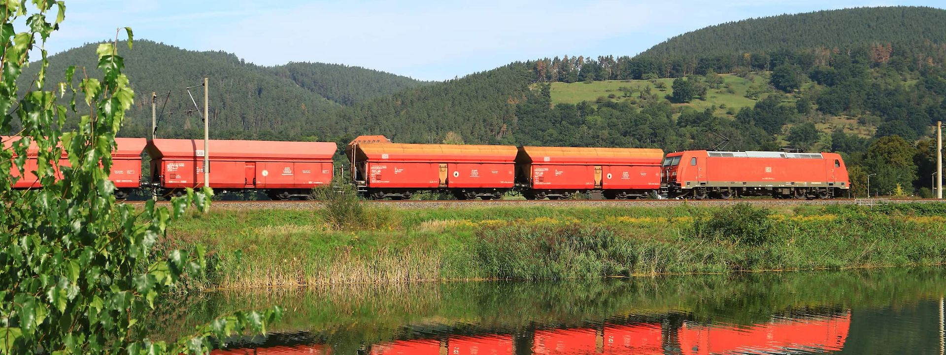 Ein Güterzug mit offenen Schüttgutwagen fährt an einem Fluss vorbei und spiegelt sich im Wasser