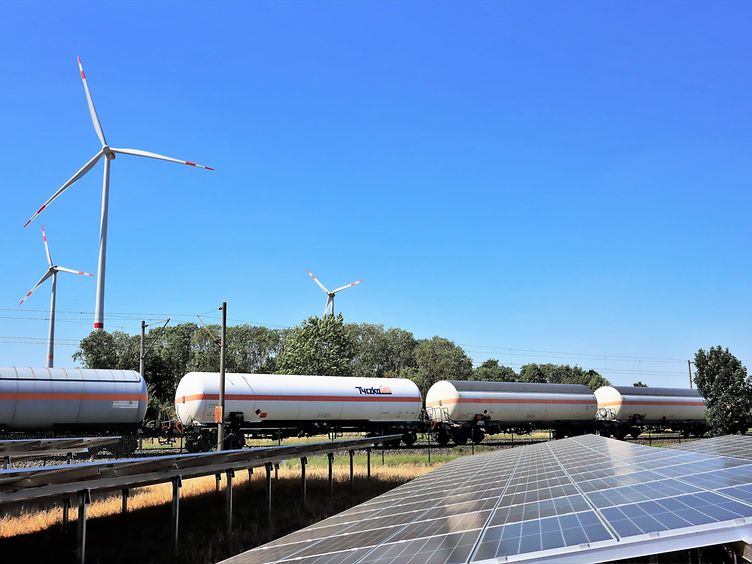 Ein Güterzug mit Druckgas-Kesselwagen fährt an einer Photovoltaikanlage vorbei, im Hintergrund sieht man Windräder