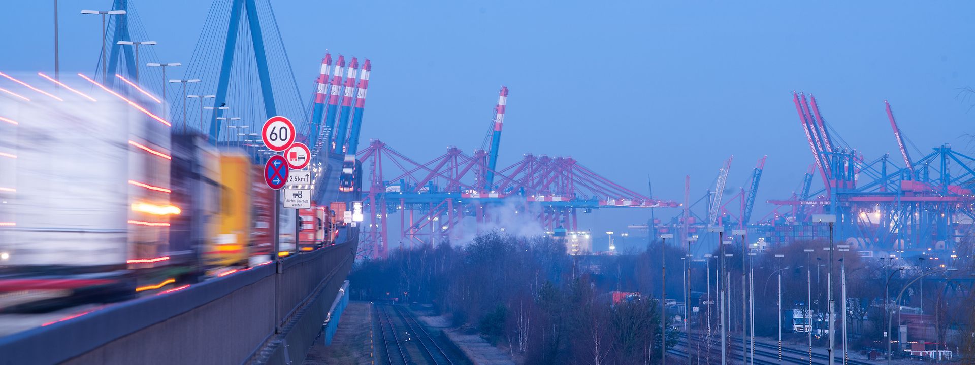 Strasse mit Lkws und Bahnschienen in Richtung Hafen mit Kränen